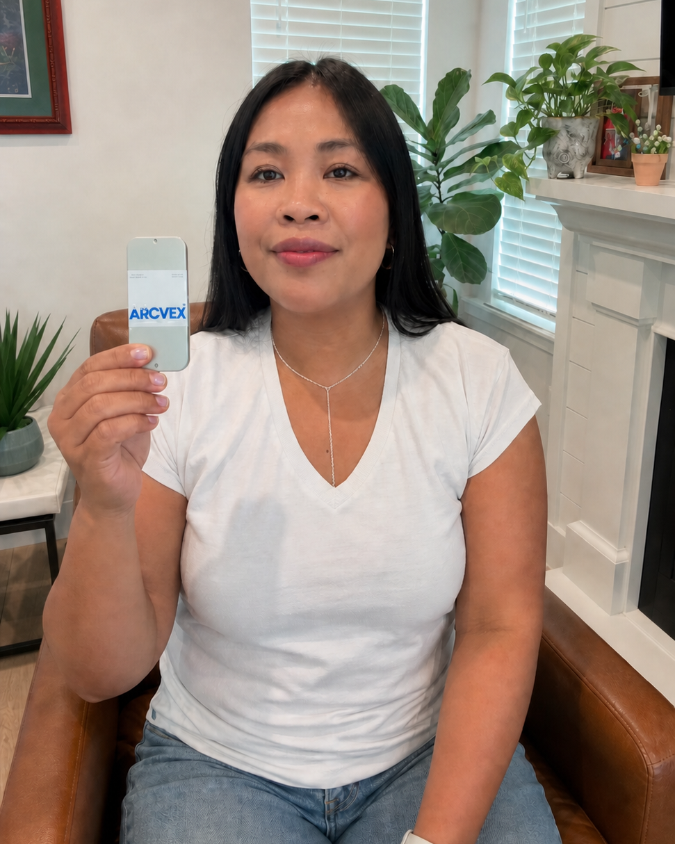 A woman sitting in a brown leather chair holds up a small white card with the brand name 'Arcvex' printed in blue. The setting is a bright, modern living room with white walls, window blinds, and indoor plants.