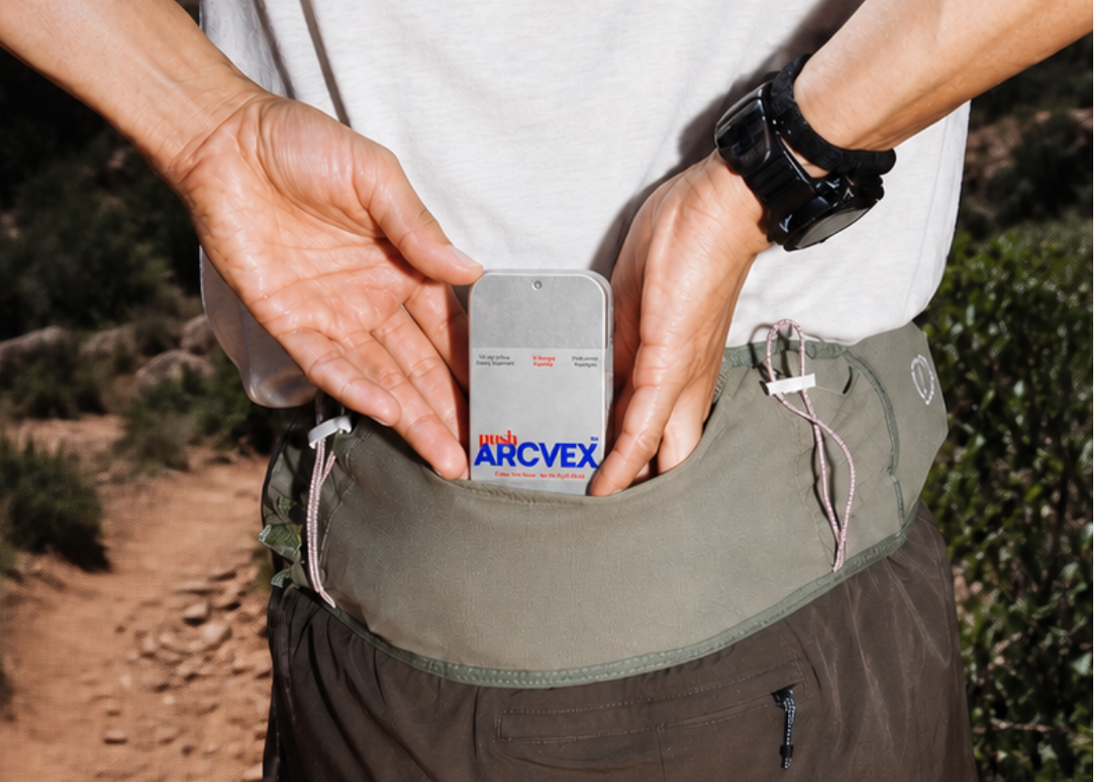 A close-up lifestyle shot of a person outdoors on a trail, showing a gray Arcvex phone pouch tucked into the back pocket of their olive green waist pack.