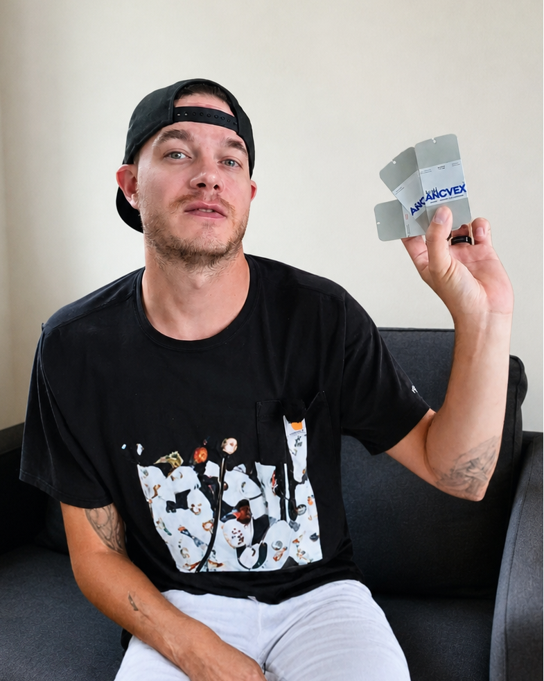 A man wearing a black graphic t-shirt and white pants sits on a dark gray couch, holding up a small gray square product with blue text that reads 'Arcvex'.